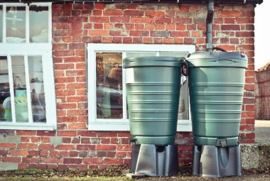 Green water barrels connected in tandem harvesting rainwater from a brick farmhouse.