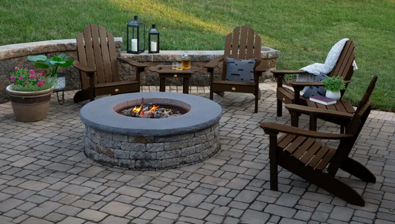 Four brown Adirondack chairs on a stone patio around a brick fire pit.