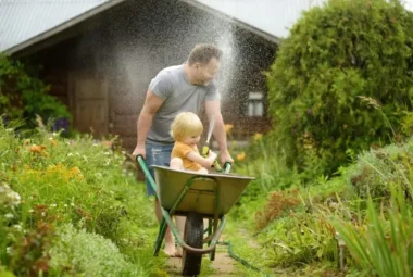 Little boy in a wheelbarrow watering dad instead of the garden.